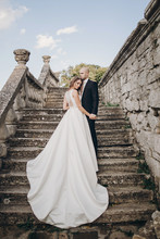 Gorgeous Wedding Couple Walking On Stone Stairs Near Old Castle In Park. Stylish Beautiful Bride In Amazing Gown And  Groom Posing On Background Of Ancient Building