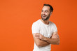 © ViDi Studio - Laughing young man in casual white t-shirt posing isolated on orange background, studio portrait. People sincere emotions lifestyle concept. Mock up copy space. Holding hands crossed, looking aside.