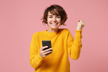 Joyful young brunette woman girl in yellow sweater posing isolated on pastel pink background. People lifestyle concept. Mock up copy space. Using mobile phone typing sms message, doing winner gesture.