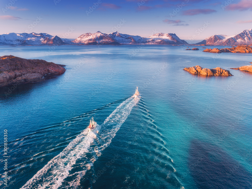 Aerial view of fishing boats, rocks in the blue sea, snowy mountains and colorful sky with clouds at sunset in winter in Lofoten islands, Norway, Landscape with two motorboats. Top view. Travel - obrazy, fototapety, plakaty 