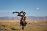 Fototapeta  - A proud young kazakh eagle hunter posing with his golden eagle on horseback on the backdrop of blue sky. Ulgii, Mongolia.