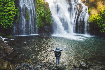 Wall Mural - Young happy woman enjoying waterfall in Bali. Wearing grey raincoat from water spray. Travel in Indonesia.