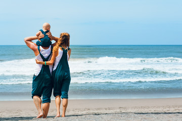 Happy family - young hipster father, mother, funny baby son walk together by tropical beach along sea surf. Little child sit on shoulders and have fun. Travel lifestyle, summer vacation with kids