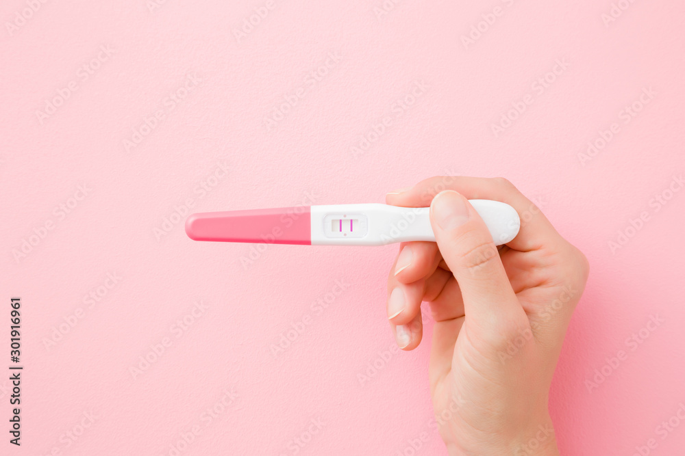 Young woman hand holding pregnancy test with two stripes on pastel pink background. Positive result. Closeup. Point of view shot. Top down view. - obrazy, fototapety, plakaty 