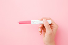 Young Woman Hand Holding Pregnancy Test With Two Stripes On Pastel Pink Background. Positive Result. Closeup. Point Of View Shot. Top Down View.