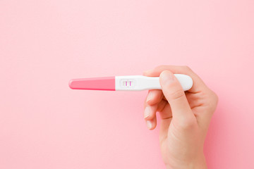 Young woman hand holding pregnancy test with two stripes on pastel pink background. Positive result. Closeup. Point of view shot. Top down view.