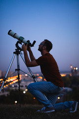 Wall Mural - astronomer with a telescope watching at the stars and moon with blurred city lights in the backgroun