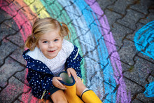 Happy Little Toddler Girl In Rubber Boots With Rainbow Sun And Clouds With Rain Painted With Colorful Chalks On Ground Or Asphalt In Summer. Cute Child Having Fun. Creative Leisure