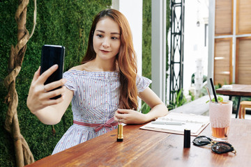 Wall Mural - Pretty young Asian woman taking selfie on smartphone after applying new brown lipstick