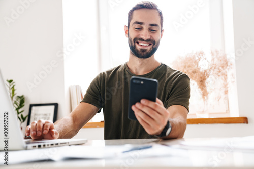 Fotografía Image of handsome happy man typing on laptop and using cellphone