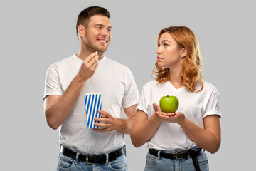 Poster - food, eating and diet concept - portrait of happy couple in white t-shirts with popcorn and green apple over grey background