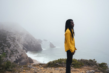 African male wearing long dreadlocks standing on ocean shore, white waves and cliffs in fog on background