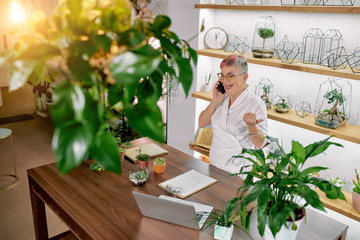 Wall Mural - Happy female in white bacthrobe, having modern hairstyle, wearing eyeglasses talk on smartphone and laugh. Laptop, papers and plants on work table