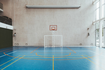 Front view of the court in the gymnasium hall; an indoor modern office stadium with a basketball basket and hoop, football goal, blue floor, a concrete wall with an undeployed projection screen above