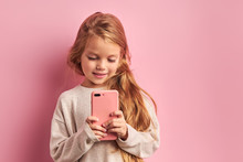 Addicted To Phone Adorable Caucasian Girl Reading And Sending Sms To Friend Isolated Over Pink Background. Mobile Phone Digital Device And Children Concept
