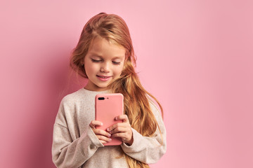 Addicted to phone adorable caucasian girl reading and sending sms to friend isolated over pink background. Mobile phone digital device and children concept