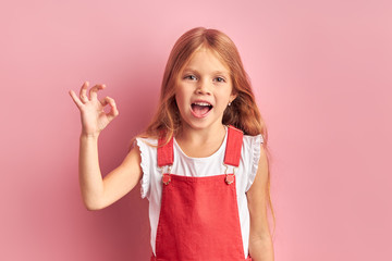 ok. little caucasian girl in red overalls show gesture ok looking at camera, smiling.