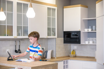 Smart preteen schoolboy doing his homework with digital tablet at home. Child using gadgets on his kitchen to study. Modern education and learning for kids