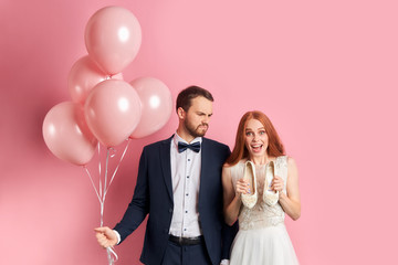 Wall Mural - Happy woman and sad struct man in suit stand isolated over pink background, holding shoes and pink air balloons