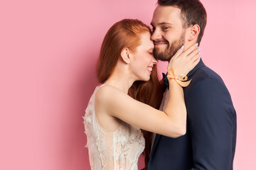 Wall Mural - Portrait of redhaired woman in white dress and bearded man in suit hugging each other, isolated pink background