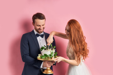 Wall Mural - Bearded man in tuxedo and woman in white dress holding sweet cake, celebrate their wedding day together