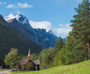 Kors Church, old wooden parish church in Rauma in Romsdal valley, Norway with road E136, green forest and mountain massif Trolltindene, Troll wall Trollveggen. Summer blue sky white clouds.