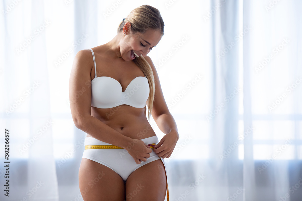 Happy young woman because she has lost weight measuring her body at home. - obrazy, fototapety, plakaty 