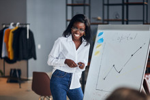 Positive African Business Lady In White Shirt And Blue Jeans Stand Talking Explaining Making Flip Chart Presentation For Younger Leaders, Share Business Experience With Smile