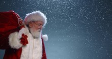 Santa Walking Through Snow, Carrying A Bag Full Of Presents, Then Stopping To Look At Camera To Playfully Wink- Christmas Spirit Concept Close Up 4k Footage