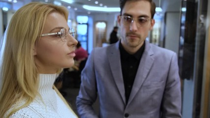 Poster - Elegant consultant is giving a pair of glasses to the pensive lady to try. She is wearing them and smiling, focus is on blond woman.