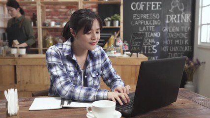 Wall Mural - charming woman freelance worker sitting at table in cafe store writing in notebook and making note. female concentrated looking at laptop computer screen. girl barista working in counter coffee shop.