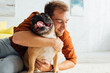 © LIGHTFIELD STUDIOS - Smiling man hugging funny French bulldog on floor in living room