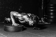 © Srdjan - Young strong sweaty focused fit muscular man with big muscles performing push ups with one hand on the barbell weight plate for cross training hard core workout in the gym  black and white