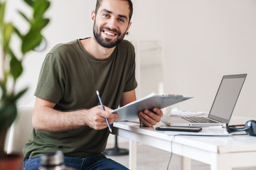 Poster - Image of smiling young man using laptop and holding clipboard