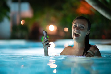 woman using mobile cell phone app on vacation in pool at night on travel holidays. Girl using smartphone app looking at screen smiling happy. Screen light on face