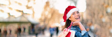 Christmas Shopping Happy Asian Girl Wearing Santa Hat Holding Gift Bags Looking At Shop In City Stores For Winter Holidays Banner Panorama Copy Space Background. Smiling Buying Presents Panoramic.