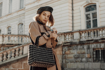 Outdoor autumn fashion portrait of young elegant woman wearing classic beige trench coat, trendy faux leather beret, holding black tweed bag, handbag, posing in street of city. Copy, empty space 