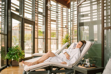 Side View Of Adult Woman In White Bathrobe Lying With Closed Eyes On Daybed After Spa Procedures In Modern Salon