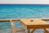 Fototapeta  - rustic patio cafe exterior of wooden table and white chairs empty furniture on Aegean sea waterfront walking district with sea surface background in summer clear weather bright day time, copy space