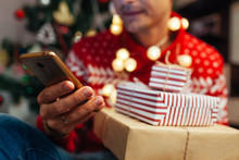 Christmas Shopping Online. Man Buying New Year Presents Using Smartphone. Guy Holding Gift Boxes And Phone By Tree