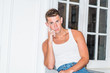 © Alexander Image - Young American Man with short hair, relaxing inside at home in New York City, wearing sleeveless, white tank top, sitting at sofa chair, by white wall, window, finger touching head, looking, thinking.