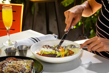 Haute cuisine - grilled sandwich with avocado guacamole sauce and two sunny side up eggs with woman hand and fork outside restaurant in a sunny day