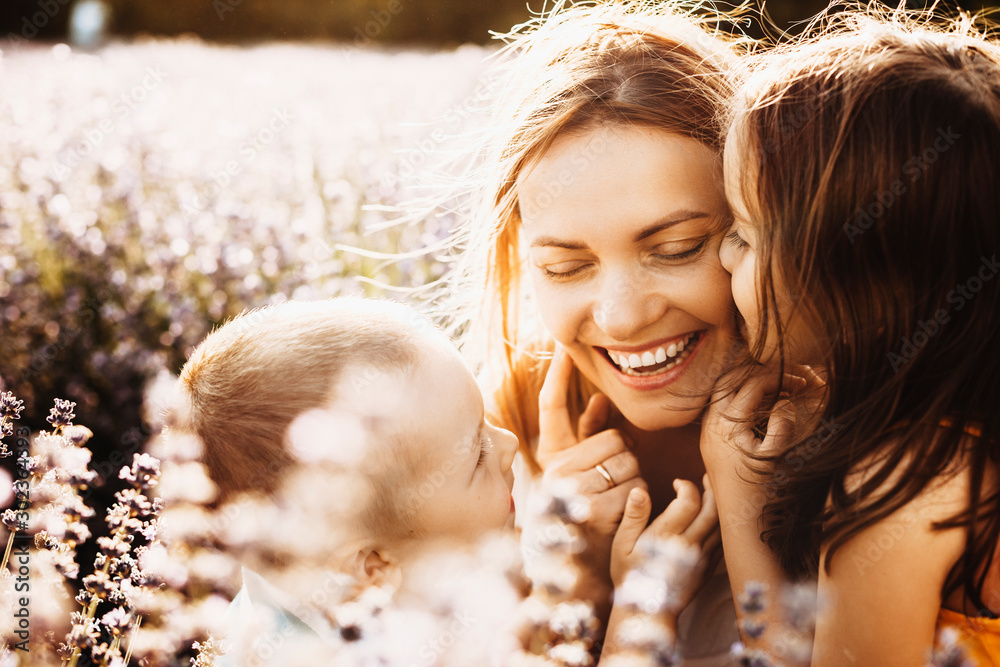 Beautiful young mother laughing while her kids are embracing and kissing her outside in a field of flowers against sunset. - obrazy, fototapety, plakaty 