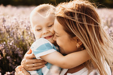 Close Up Portrait Of A Young Mother Embracing Her Son With Eyes Closed Smiling Outdoor In Field Of Flowers Against Sunset.
