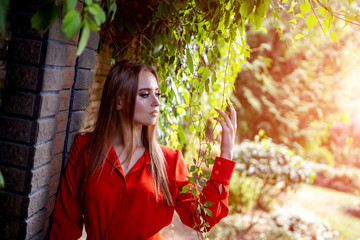 Wall Mural - Photo of attractive woman in red dress standing near the brick wall. Park view. Spring, autumn. Young girl in bright light and green leaves. Blurred background. Closeup.