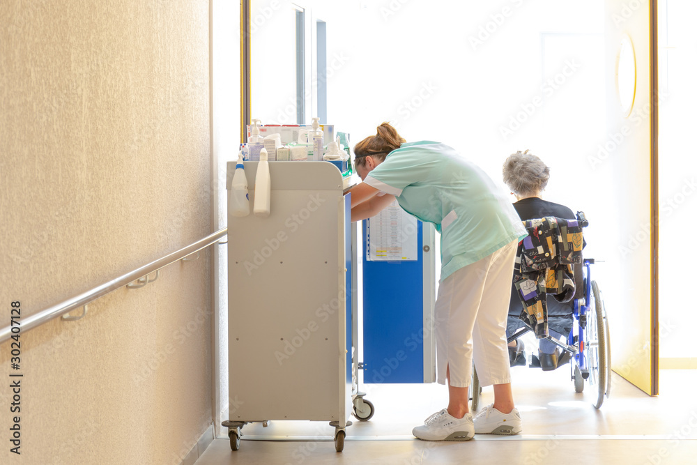 Aide soignante au travail accompagne personne agée en fauteuil roulant dans couloir hopital clinique maison de retraite - obrazy, fototapety, plakaty 