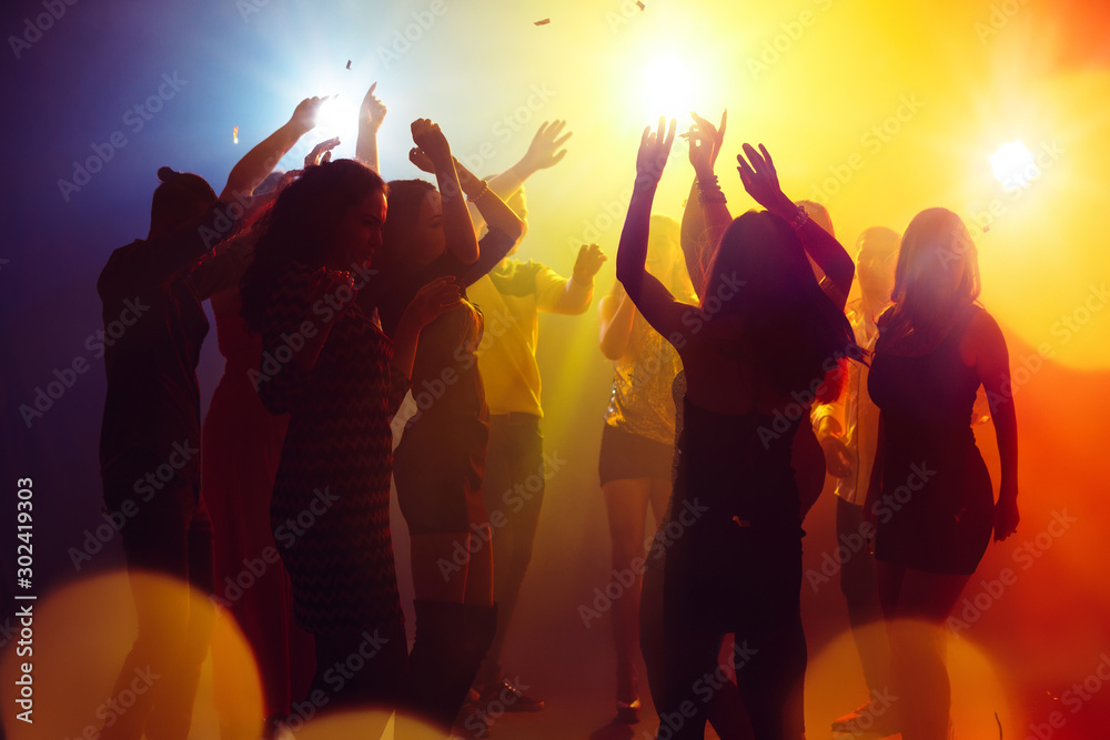 Holidays. A crowd of people in silhouette raises their hands on dancefloor on neon light background. Night life, club, music, dance, motion, youth. Yellow-blue colors and moving girls and boys. - obrazy, fototapety, plakaty 