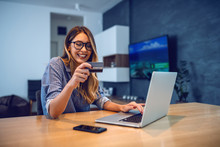Young Attractive Smiling Caucasian Woman With Eyeglasses Holding Credit Card And Typing Number Of Her Bank Account On Laptop While Sitting At Dining Table. Online Shopping Concept.