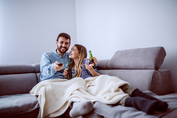 Cute caucasian couple in love sitting on sofa in living room,drinking beer and watching tv. Man holding remote control and changing channel.