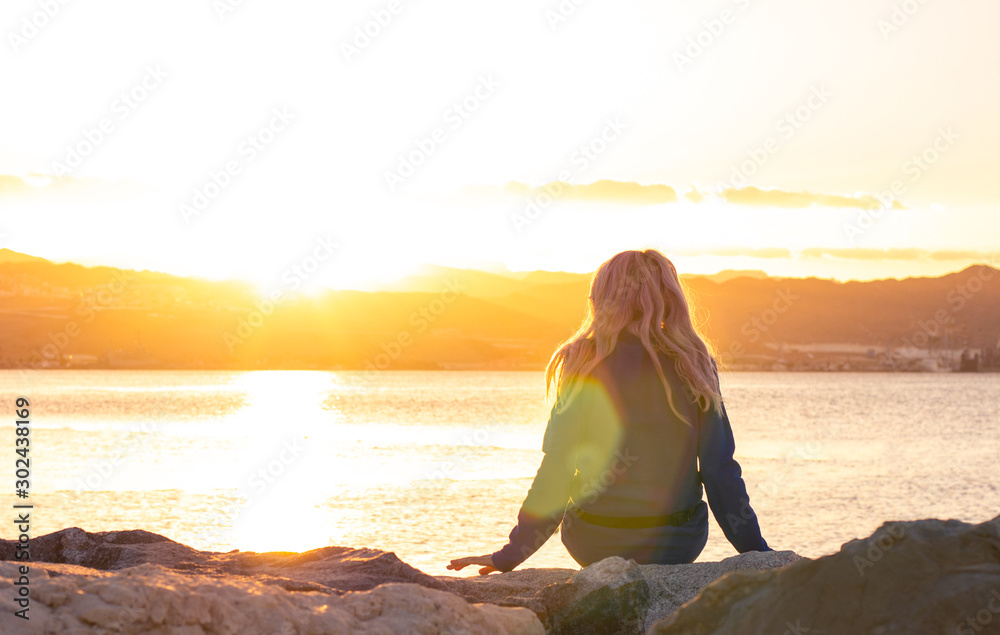 lonely girl sitting back to camera on stone sea bay waterfront shore line and enjoy by beautiful bright orange sunset light with colorful glares , thinking and human emotion concept photography  - obrazy, fototapety, plakaty 